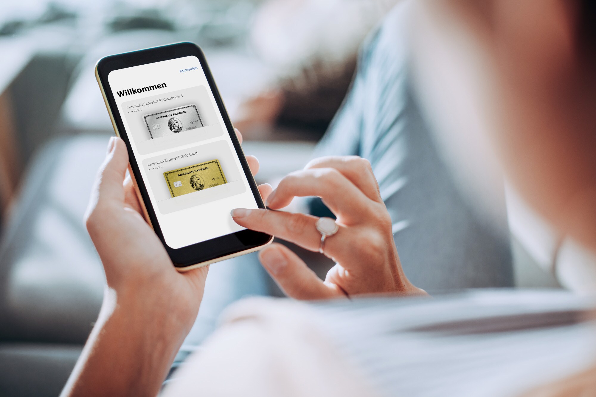 Nahaufnahme eines Smartphones mit der Amex App auf dem Display, in den Händen einer Frau auf einem Sofa